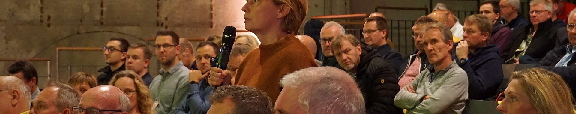 Eine Frau steht mit Mikrofon in einer sitzenden Zuschauermenge und spricht. 