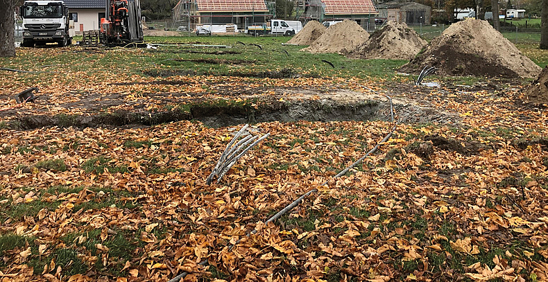 Erdkabeln auf einem mit Herbstlaub bedeckten Gelände. Im Hintergrund sind Rohbauten mit Gerüsten und Fahrzeugen zu sehen.