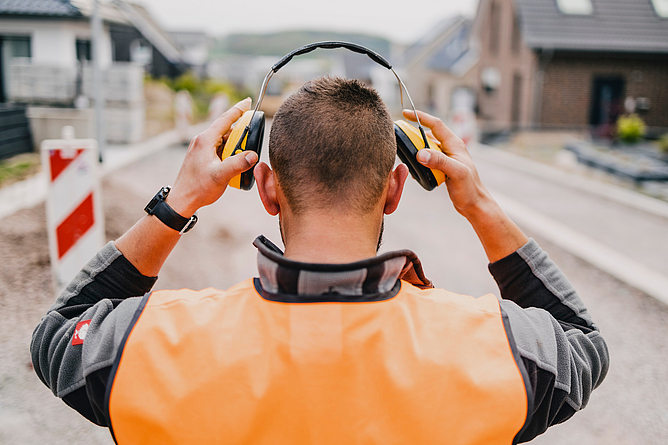 Blick auf einen Bauarbeiter von hinten, der seinen Gehörschutz aufsetzt.