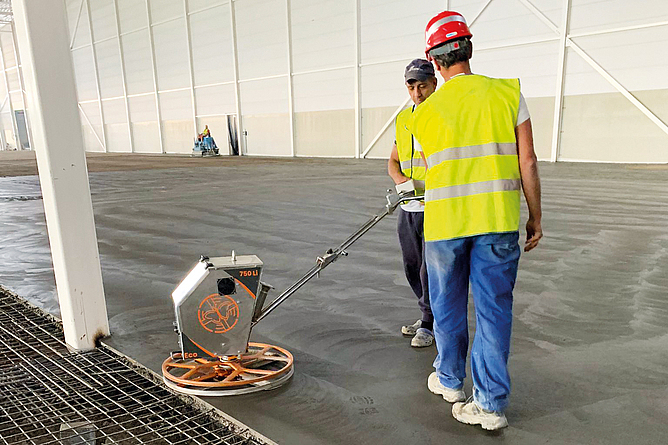 Zwei Männer mit Schutzhelmen und Warnweste an einem Einfachglätter am Rande einer Betonfläche