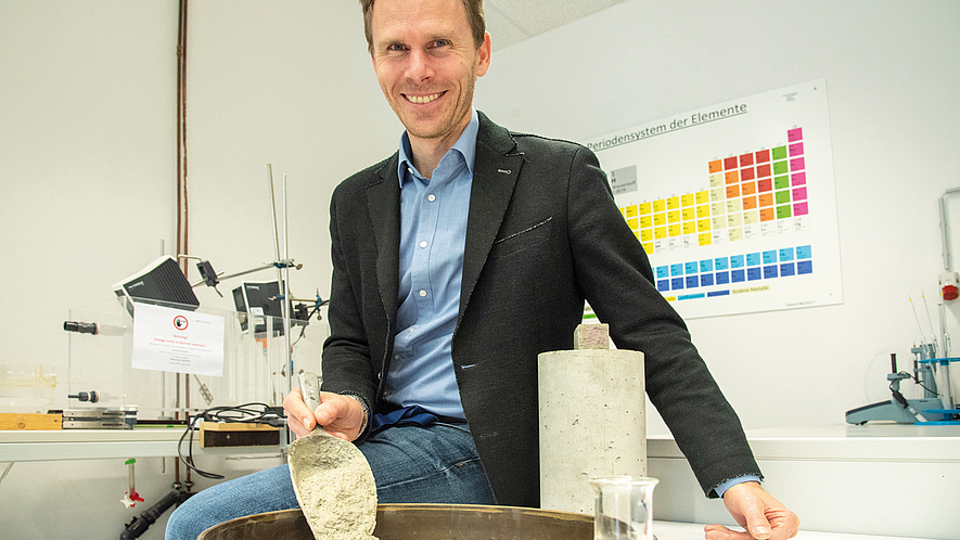 Ein lächelnder Mann sitzt in einem Labor mit einem Bein auf einem Tisch, vor ihm auf dem Tisch steht ein Schale mit Gesteinsmaterialien und Zement, daneben eine Glasflasche.