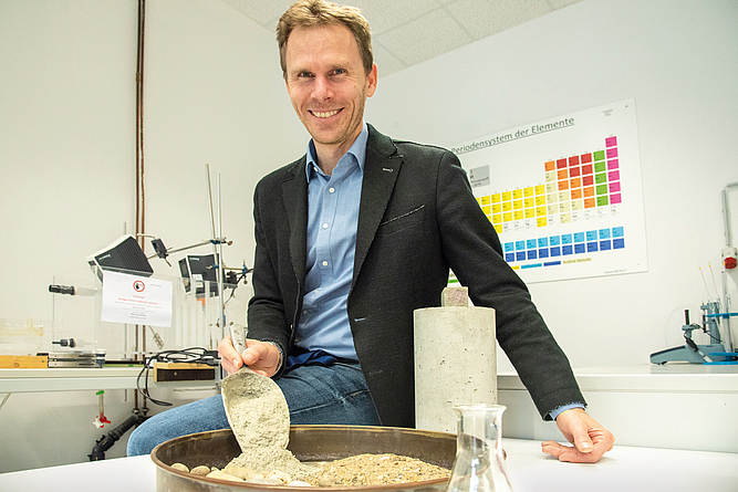 Ein lächelnder Mann sitzt in einem Labor mit einem Bein auf einem Tisch, vor ihm auf dem Tisch steht ein Schale mit Gesteinsmaterialien und Zement, daneben eine Glasflasche.