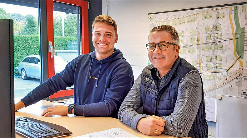 Ein jüngerer und ein älterer Mann stehen lächelnd nebeneinander an einem Tisch, auf dem sich ein Monitor und eine Tastatur befinden.