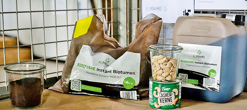 Das von B2Square hergestellte Biobitumen enthält u. a. ein Extrakt aus gepressten Cashewnussschalen. Ein Beutel "B2Square Instant Biotumen" steht neben einer Packung Cashew-Kerne, auf der sich ein Glas mit Cashew-Nüssen befindet.