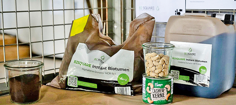 Ein Beutel "B2Square Instant Biotumen" steht neben einer Packung Cashew-Kerne, auf der sich ein Glas mit Cashew-Nüssen befindet.