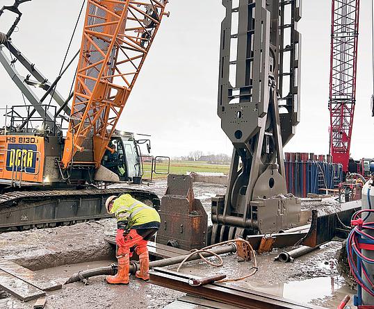 Nahaufnahme eines Seilbaggers mit Schlitzwandgreifer, der in einer rechteckigen Öffnung versinkt. Daneben bückt sich eine Person mit Warnkleidung über die Öffnung. 