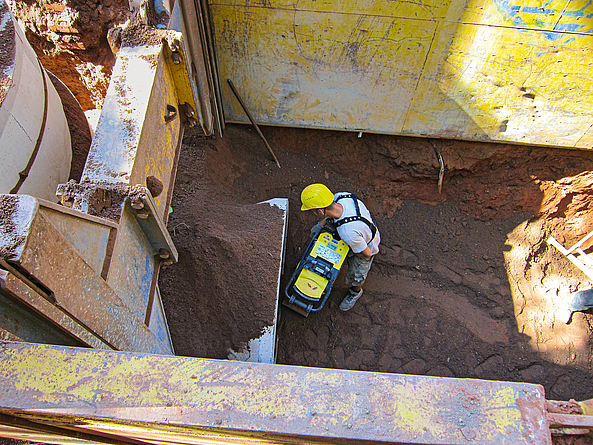 Blick von oben in einen Graben, in dessen Sohle ein Mann mit Helm dunkle Erde mit einem Geräte verdichtet.