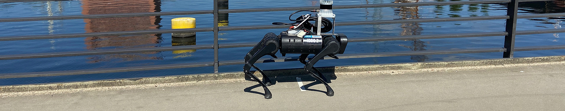 Roboter in Tierform mit vier beweglichen Roboterbeinen. Der Roboter bewegt sich auf einer Brücke, die über ein Gewässer führt. 