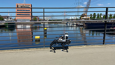Roboter in Tierform mit vier beweglichen Roboterbeinen. Der Roboter bewegt sich auf einer Brücke, die über ein Gewässer führt. 