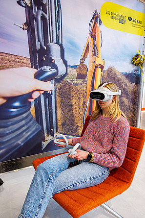 Besucherin bei der VR-Anwendung zum Anschnallen in Baumaschinen auf der bauma 2025 Frau im Sessel testet VR-Brille. Im Hintergrund ein Bild eines Baggers aus der Sicht des Baggerfahrers.