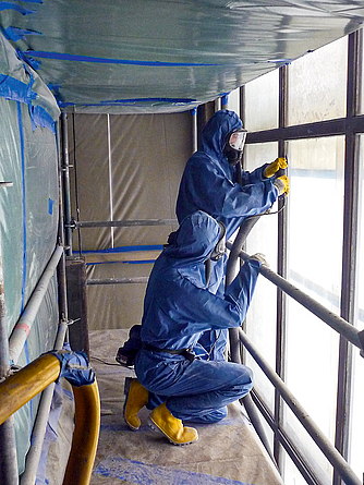 Zwei Männer in Schutzanzügen und Atemschutzmasken dichten die Fensterkonstruktion mit Fugenmasse ab.