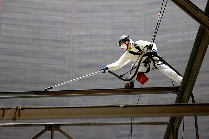 Eine Person in weißem Schutzanzug, Atem- und Kopfschutz ist mit einem Seil gesichert und reinigt mit einem Schlauch das Tragwerk.