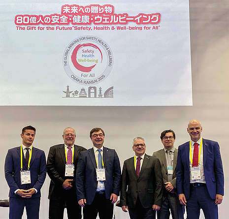 Sechs Männer in Anzügen stehen vor einer Leinwand mit dem Logo und Motto des Weltkongresses für Sicherheit und Gesundheit bei der Arbeit 2025 in Osaka.