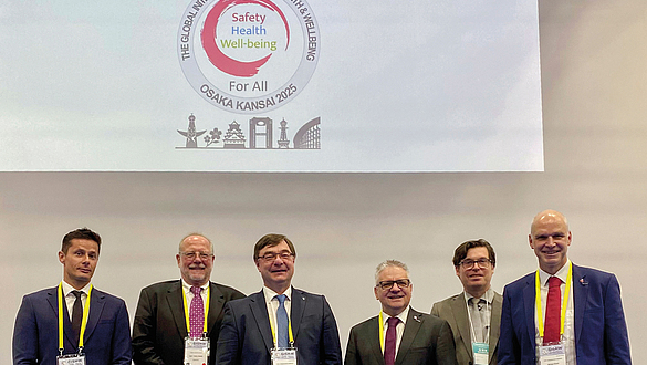 Sechs Männer in Anzügen stehen vor einer Leinwand mit dem Logo und Motto des Weltkongresses für Sicherheit und Gesundheit bei der Arbeit 2025 in Osaka.