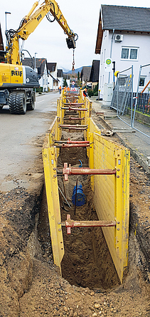 Ein Großteil der neuen Trinkwasser-Transportleitung wird in konventioneller Bauweise hergestellt. Schmaler aufgebaggerter Graben am Straßenrand eines Ortes. Gelbe vertikale Stützplatte an jeder Seite sichern das Einfallen des Grabens, dazwischen befindet sich ein halb eingegrabenes blaues Rohr.