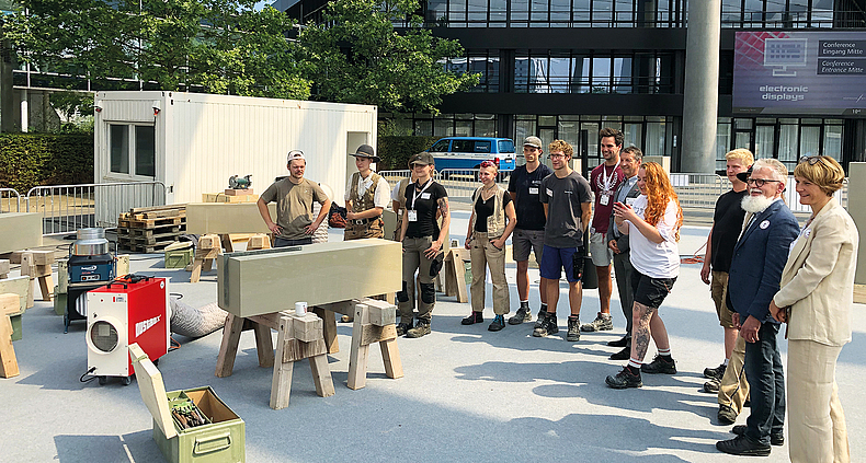 Auch beim Steinmetz-Nachwuchswettbewerb war das Thema Staubvermeidung präsent (hier ein Gruppenbild zu Beginn der Veranstaltung). Gruppenbild mit Steinmetzen, die im Halbkreis um einen aufgebockten Steinklotz stehen. Neben dem Klotz ist ein Baustauber zu sehen.