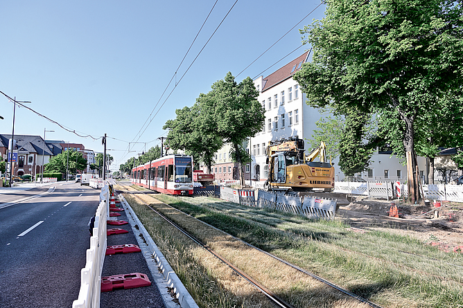 Eine Straßenbahn fährt entlang der neu gebauten Trasse neben einer Straße. Hinter einem Bauzaun steht noch ein Baufahrzeug.
