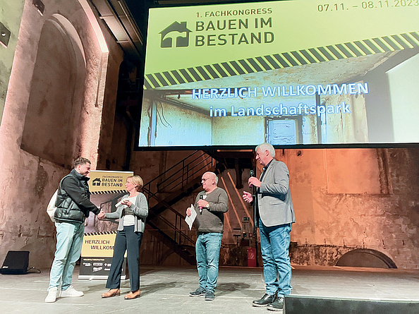 Andrea Bonner (2. v. l.) überreicht dem Gewinner (1. v. l.) des Azubi-Quiz das gewonnene Smartphone. Gerhard Cittrich (3. v. l.) von der IG BAU und Norbert Kluger von der BG BAU moderierten die Preisverleihung. Preisübergabe an den Gewinner des Azubi-Quiz durch Andrea Bonner. Mit auf der Bühne die beiden Moderatoren.