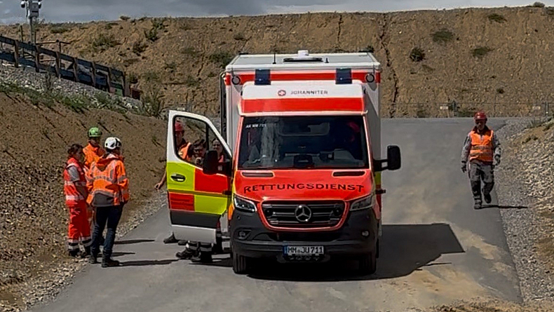 Rettungswagen mit geöffneter Beifahrertür wird an einer Baustelle von Personen mit Schutzkleidung empfangen.
