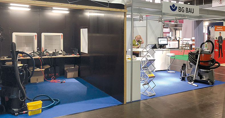 Stand der BG BAU auf der Stone+tec mit der Absaugkabine der BG BAU (Aufnahme vor Messebeginn) Erleuchteter Messestand mit dem Logo und Schriftzug der BG BAU, mit Broschürenständer, Tresen und Absauggerät, links daneben Absaugkabine.