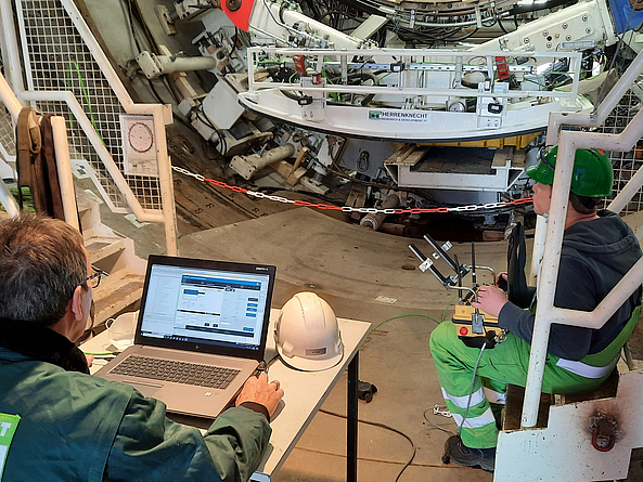 Der untere Teil einer Tunnelbohrmaschine befindet sich in einem engem Raum. Davor sitzt ein Mann am Tisch und vergleicht Daten im Laptop. Eine andere Person sitzt seitlich und steuert etwas. 
