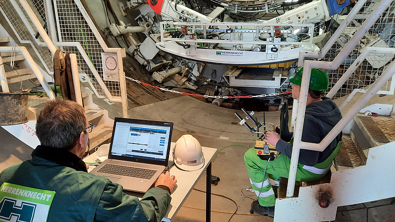 Der untere Teil einer Tunnelbohrmaschine befindet sich in einem engem Raum. Davor sitzt ein Mann am Tisch und vergleicht Daten im Laptop. Eine andere Person sitzt seitlich und steuert etwas. 