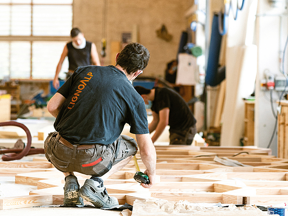 Drei Männer arbeiten in einer großen Werkstatt an einer großflächigen Holzkonstruktion.