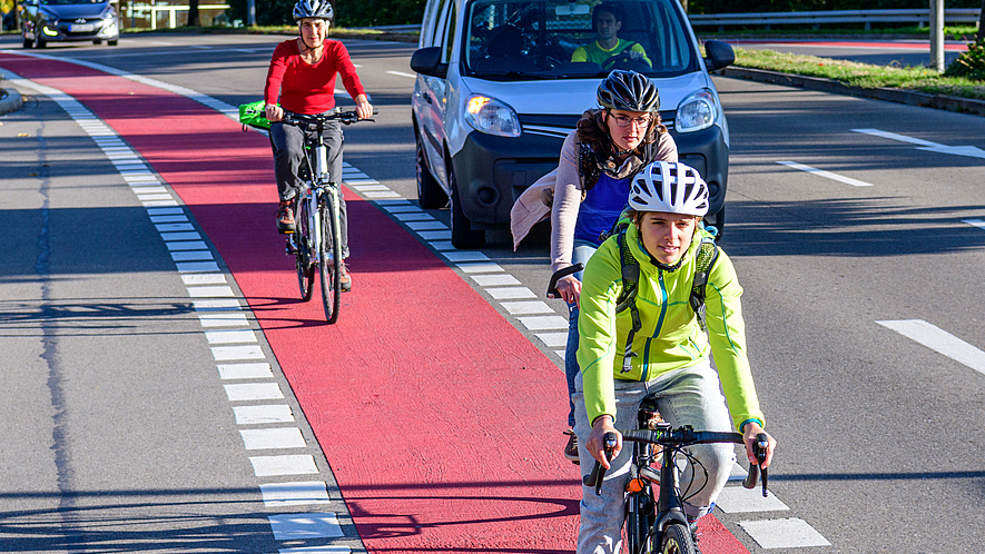 Drei Fahrradfahrer mit Helm fahren auf einem Radweg. Ein wartendes Auto möchte rechts abbiegen. 