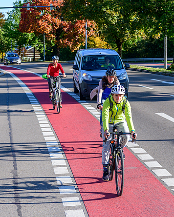 Drei Fahrradfahrer mit Helm fahren auf einem Radweg. Ein wartendes Auto möchte rechts abbiegen. 