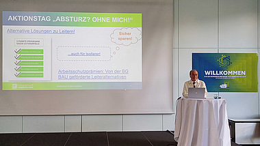 Bernd Merz steht an einem Rednertisch auf einem Podium. Vor ihm ist ein Laptop geöffnet, auf der Leinwand hinter ihm ist eine Folie zum Thema Absturz zu sehen.