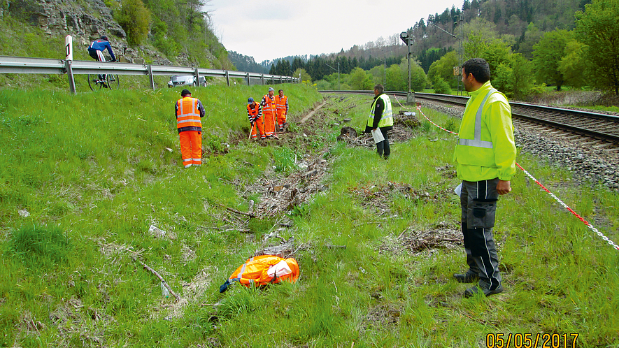 Rot-weiße Kette als sichtbare Abgrenzung Gleisarbeiter mit oranger Schutzkleidung und Sicherungsposten mit neongelber Schutzkleidung befinden sich einem mit rot-weißer Kette etwas 2 m abgesperrten Bereich zwischen Gleis und Leitplanke einer Straße.