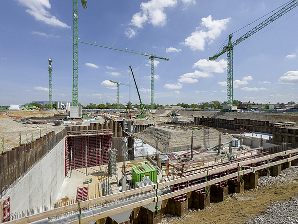 Große verbaute Baugrube, in der Rohbauarbeiten stattfinden. Um die Baugrube herum stehen Kräne.
