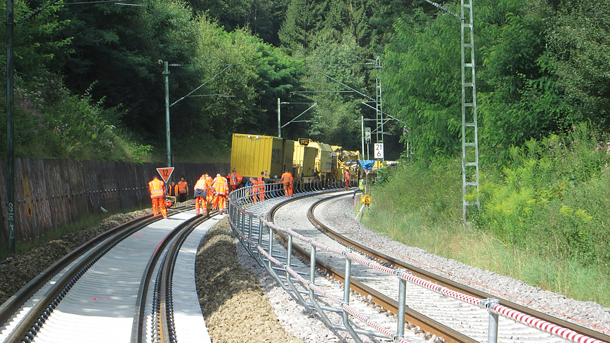 Mehrere Bahnarbeiter in orangefarbener Schutzkleidung arbeiten mit Gleisbaumaschinen an einer zweigleisigen Bahnstrecke in einer bewaldeten Kurve.