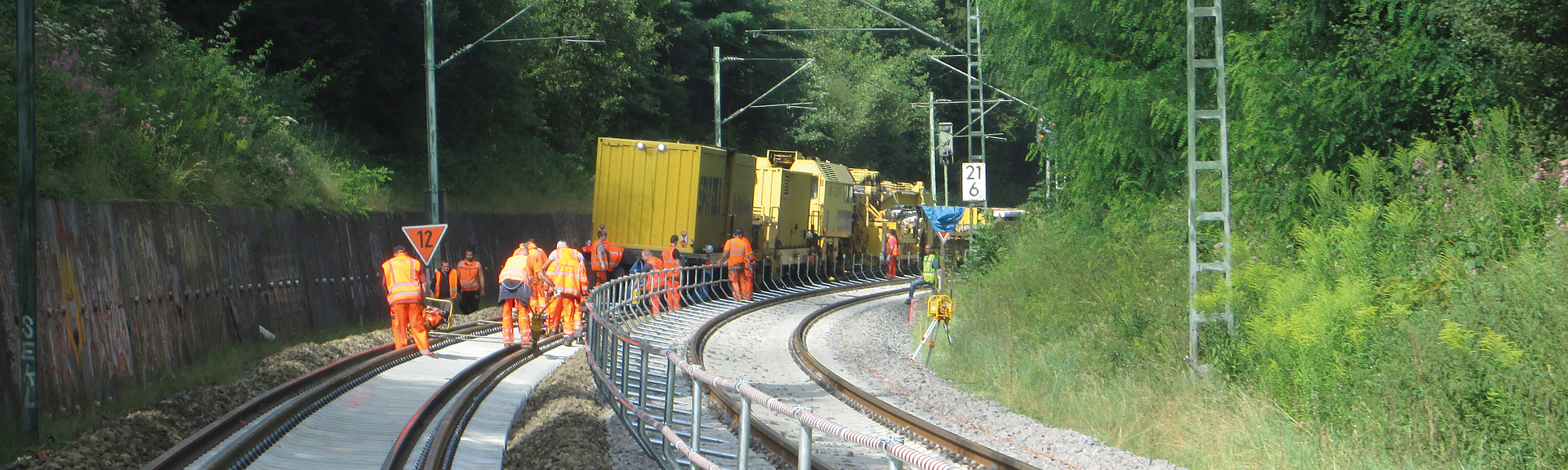 Mehrere Bahnarbeiter in orangefarbener Schutzkleidung arbeiten mit Gleisbaumaschinen an einer zweigleisigen Bahnstrecke in einer bewaldeten Kurve.