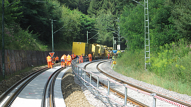 Mehrere Bahnarbeiter in orangefarbener Schutzkleidung arbeiten mit Gleisbaumaschinen an einer zweigleisigen Bahnstrecke in einer bewaldeten Kurve.