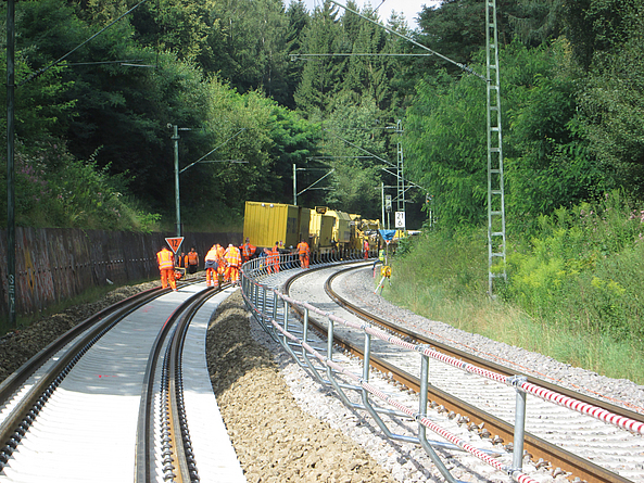 Mehrere Bahnarbeiter in orangefarbener Schutzkleidung arbeiten mit Gleisbaumaschinen an einer zweigleisigen Bahnstrecke in einer bewaldeten Kurve.