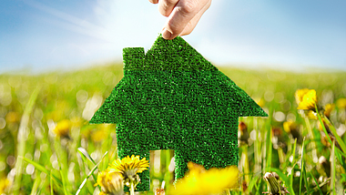 Eine Hand hält eine grün bepflanzte Hausschablone über eine Wiese bei blauem Himmel mit Sonnenschein.