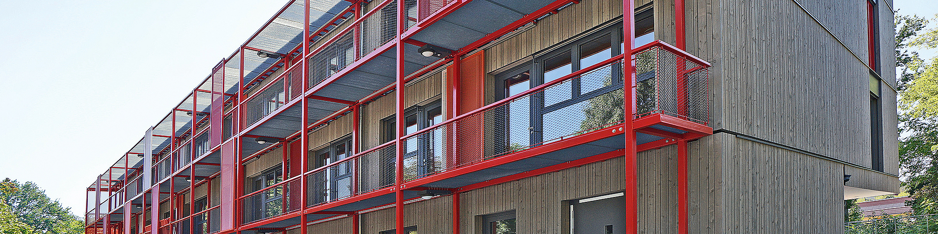 An der Fassade des Holzmodul-Neubaus einer Kita sind in jeder der zwei oberen Etagen je ein Metall-Laubengang in leuchtendem Rot zu sehen. Sie dienen nicht nur als Verbin­dungsgang vor den Fenstern, sondern auch als als zweiter Fluchtweg: Durchgehende Eckpfosten führen bis auf den Boden.