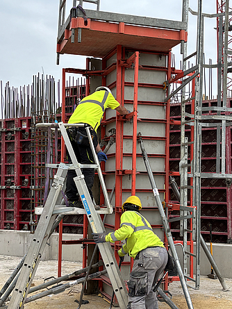 Zwei Bauarbeiter in Warnkleidung arbeiten an einer Schalungskonstruktion. Einer steht auf einer Podestleiter und befestigt Teile an einer Betonsäule, der andere unterstützt ihn vom Boden aus.