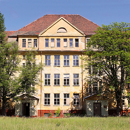 Blick auf die Vorderfront des Gebäudes mit kaputten Fenstern und einer teilweise zerstörten Fassade.