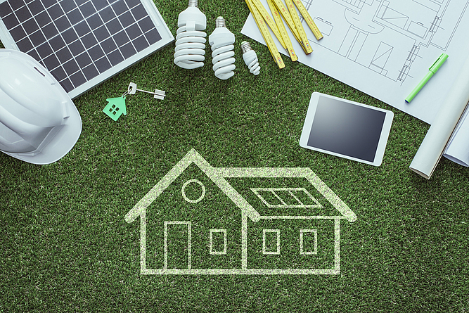 Auf einer grünen Kunstrasenfläche ist ein Wohnhaus gezeichnet. Ein Smartphone, Schlüssel und eine Bauzeichnung sind darauf abgelegt.