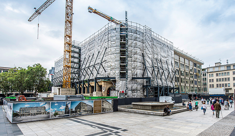 Blick von außen auf die Baustelle mit dem eingerüsteten Blau-Gold-Haus und der Bauwand mit Visualisierung des fertigen Gebäudes. Im Vordegrund laufen Passanten