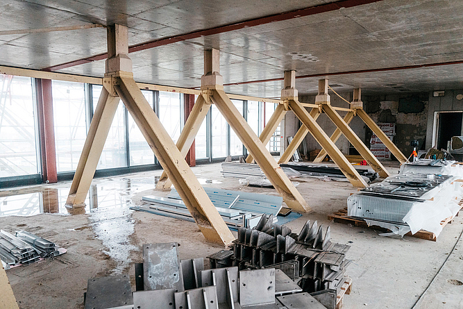 Innenraum einer Baustelle mit schrägen Stützen unter einer Betondecke und gelagerten Bauteilen.