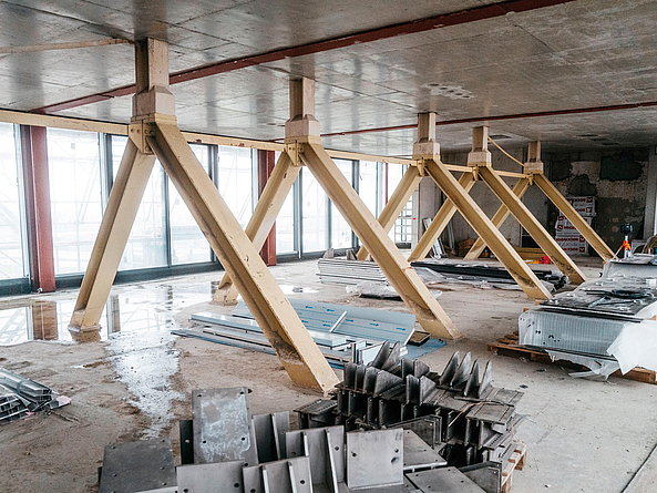 Innenraum einer Baustelle mit schrägen Stützen unter einer Betondecke und gelagerten Bauteilen.