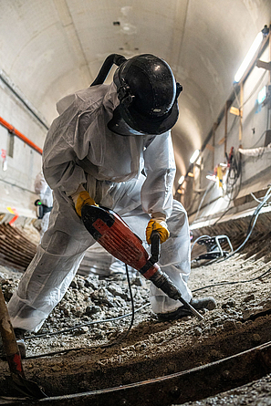 Bauarbeiter in Vollschutzkleidung (gebläseunterstützter Helm mit Partikelfilter und Schutzanzug Kategorie III Typ 5 sowie Nitrilhandschuhe) bei Abbrucharbeiten mit einem Presslufthammer im Tunnelinneren.