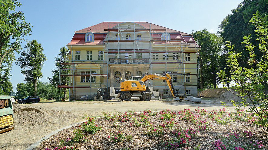 Historisches Gebäude mit Gerüst und Bagger auf einer Baustelle, im Vordergrund Blumenbeet.