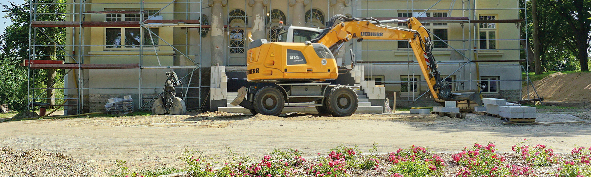 Historisches Gebäude mit Gerüst und Bagger auf einer Baustelle, im Vordergrund Blumenbeet.