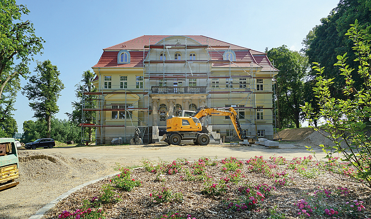 Historisches Gebäude mit Gerüst und Bagger auf einer Baustelle, im Vordergrund Blumenbeet.