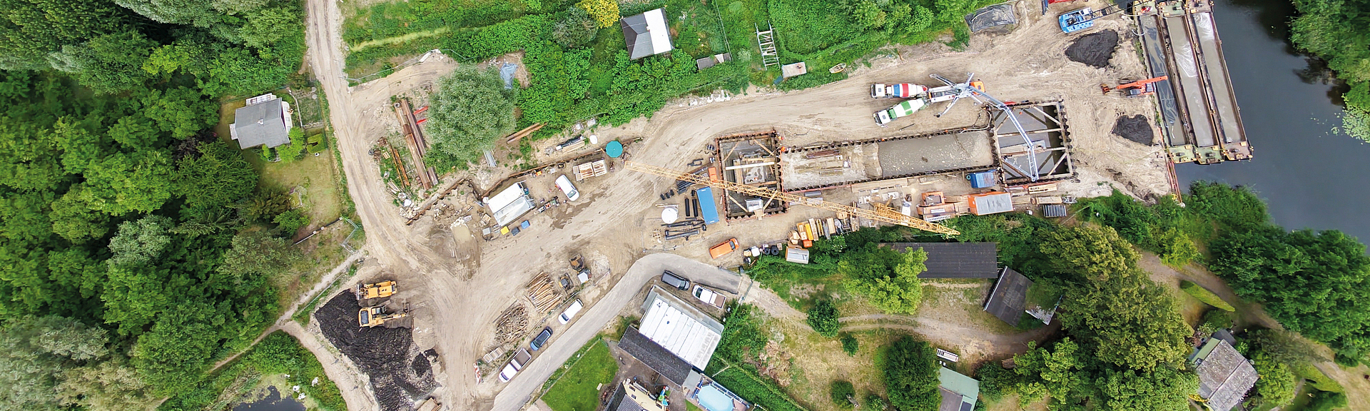 Luftaufnahme einer Brückenbaustelle mit Baumaschinen und Baumaterial zwischen einem Fluss und angrenzenden Wohngebäuden.