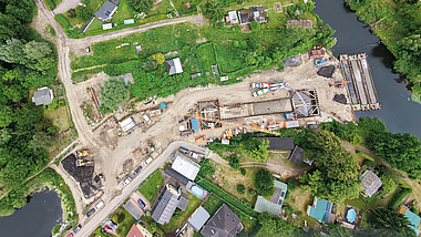 Luftaufnahme einer Brückenbaustelle mit Baumaschinen und Baumaterial zwischen einem Fluss und angrenzenden Wohngebäuden.